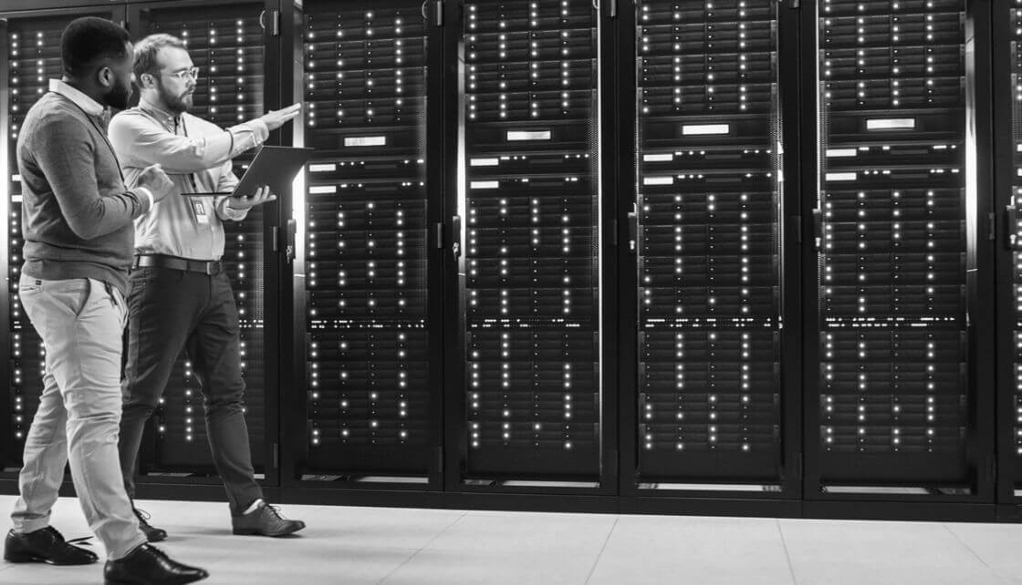Two men using a laptop standing near multiple servers